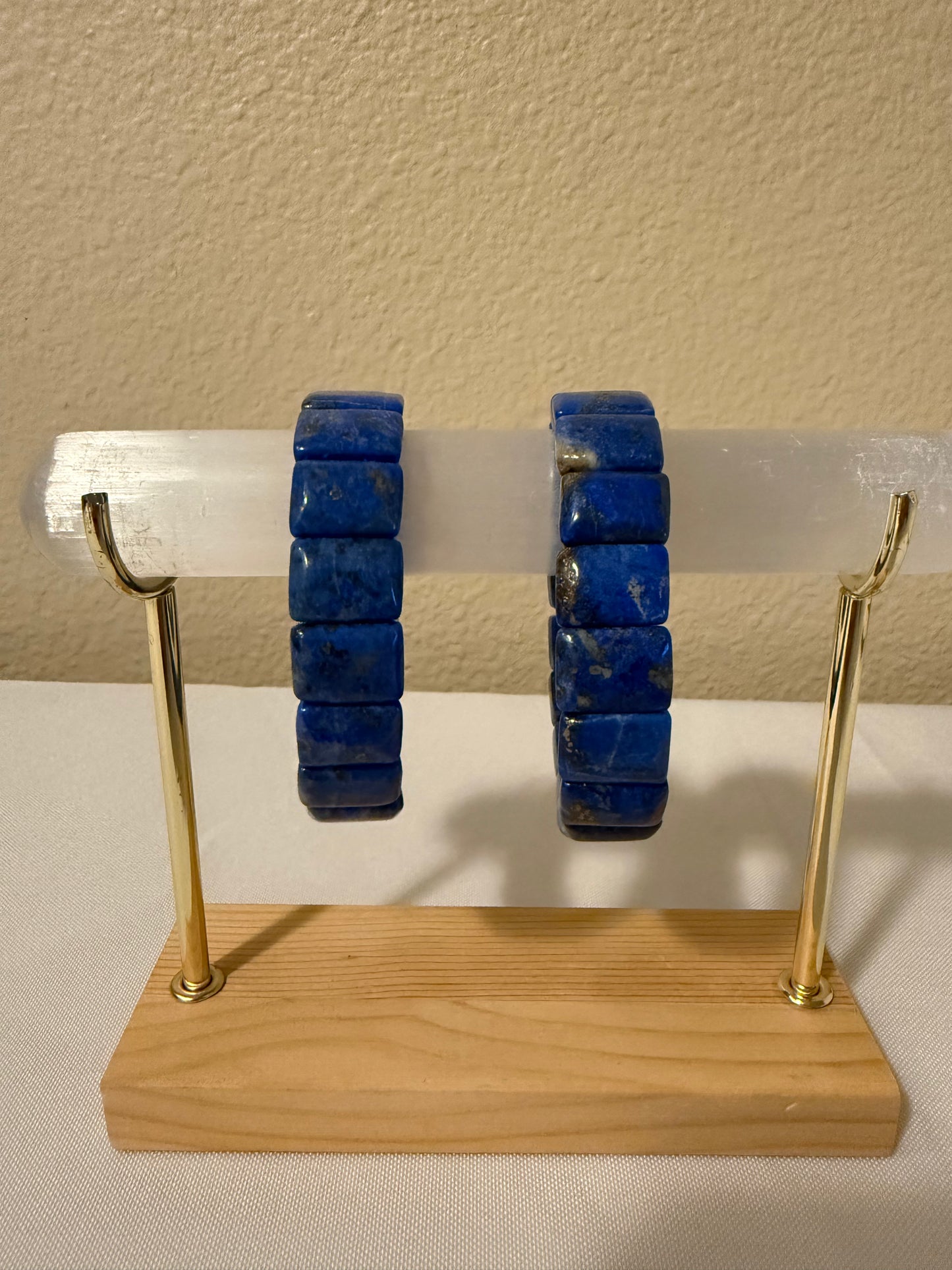 Two lapiz lazuli bangles displayed on a clear acrylic rod mounted on a wooden stand with gold-colored metal arms, set against a beige wall and white surface.