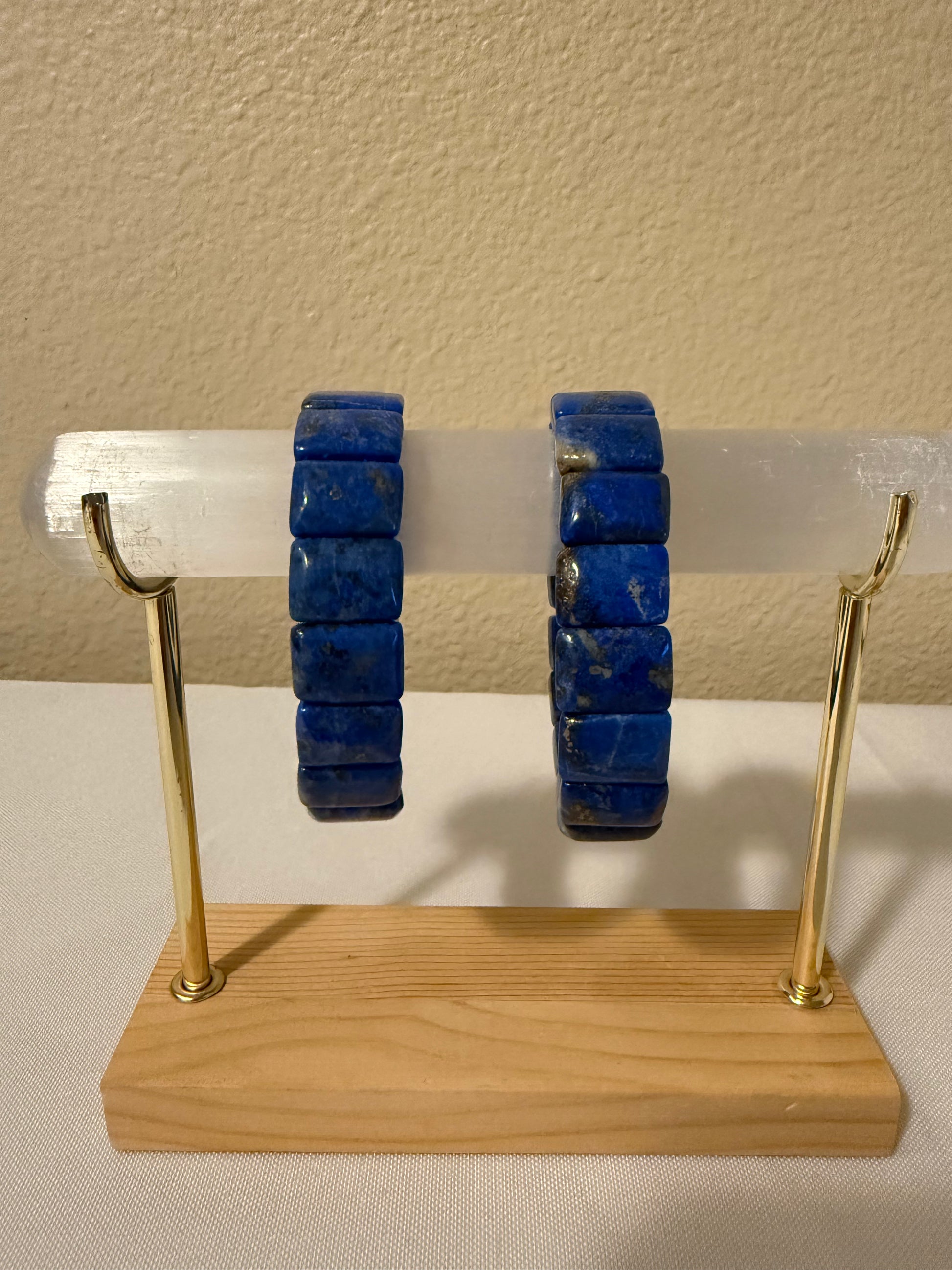Two lapiz lazuli bangles displayed on a clear acrylic rod mounted on a wooden stand with gold-colored metal arms, set against a beige wall and white surface.