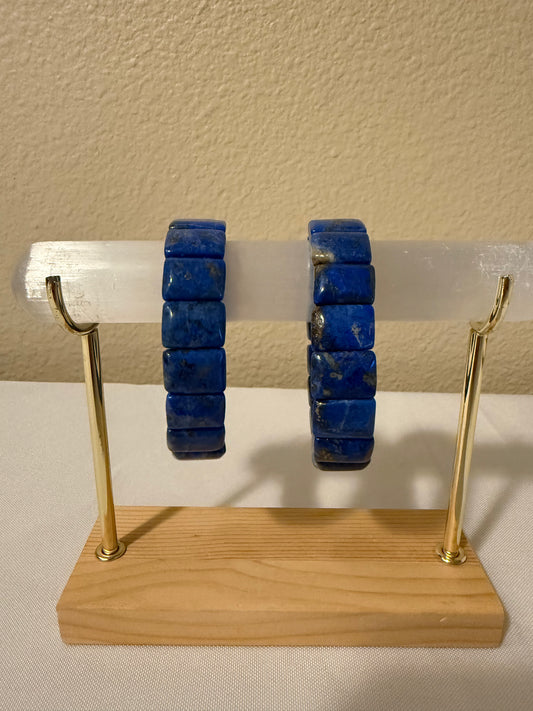 Two lapiz lazuli bangles displayed on a clear acrylic rod mounted on a wooden stand with gold-colored metal arms, set against a beige wall and white surface.