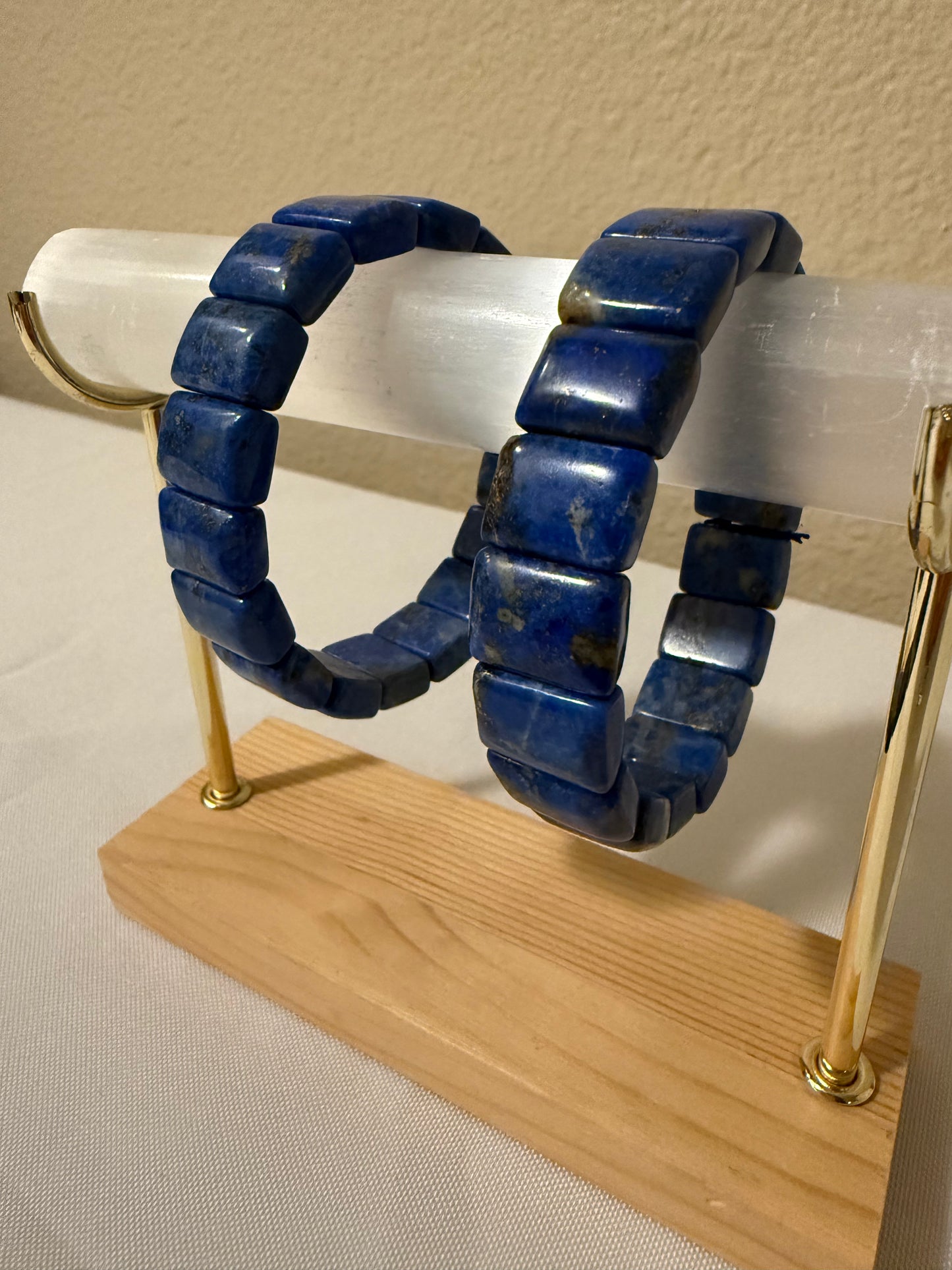 Two blue lapis lazuli bead bracelets displayed on a clear acrylic stand with a wooden base and gold-tone metal supports.