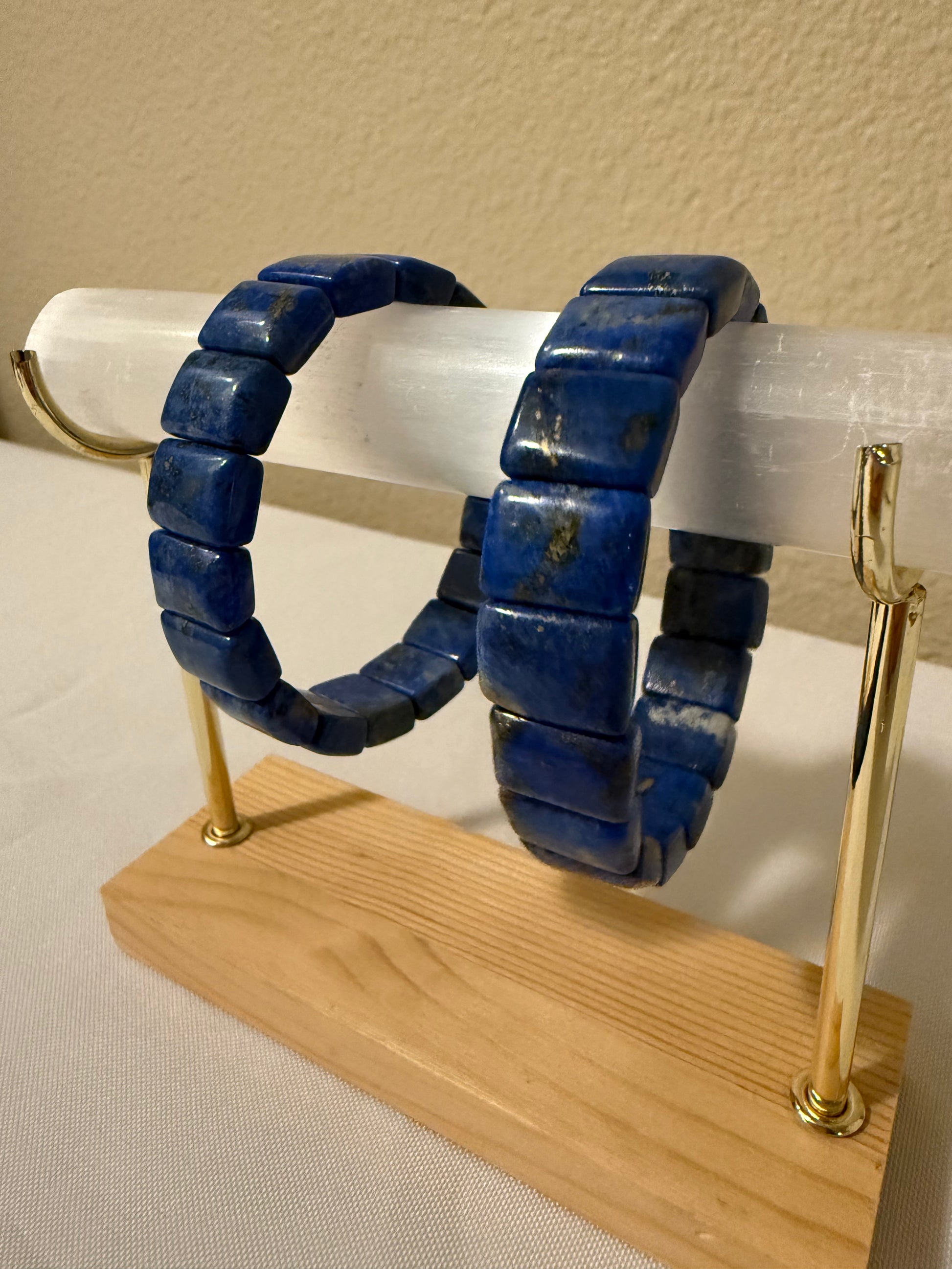 A pair of lapiz lazuli beaded bracelets displayed on a clear acrylic stand with a wooden base and gold-tone metal supports against a beige wall.